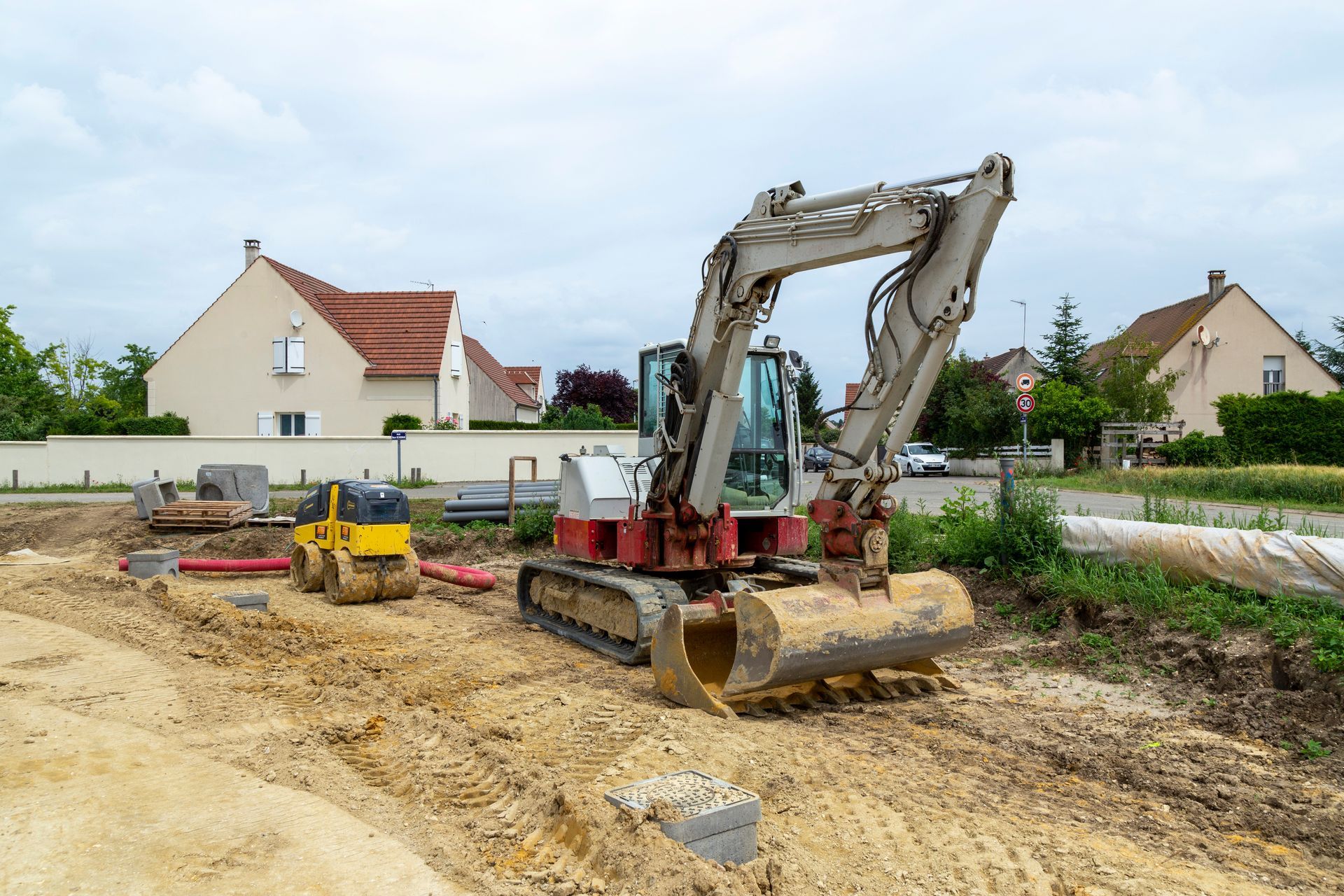 Une pelleteuse sur un chantier de construction en terre près de maisons, avec une petite machine jaune et des arbres en arrière-plan.