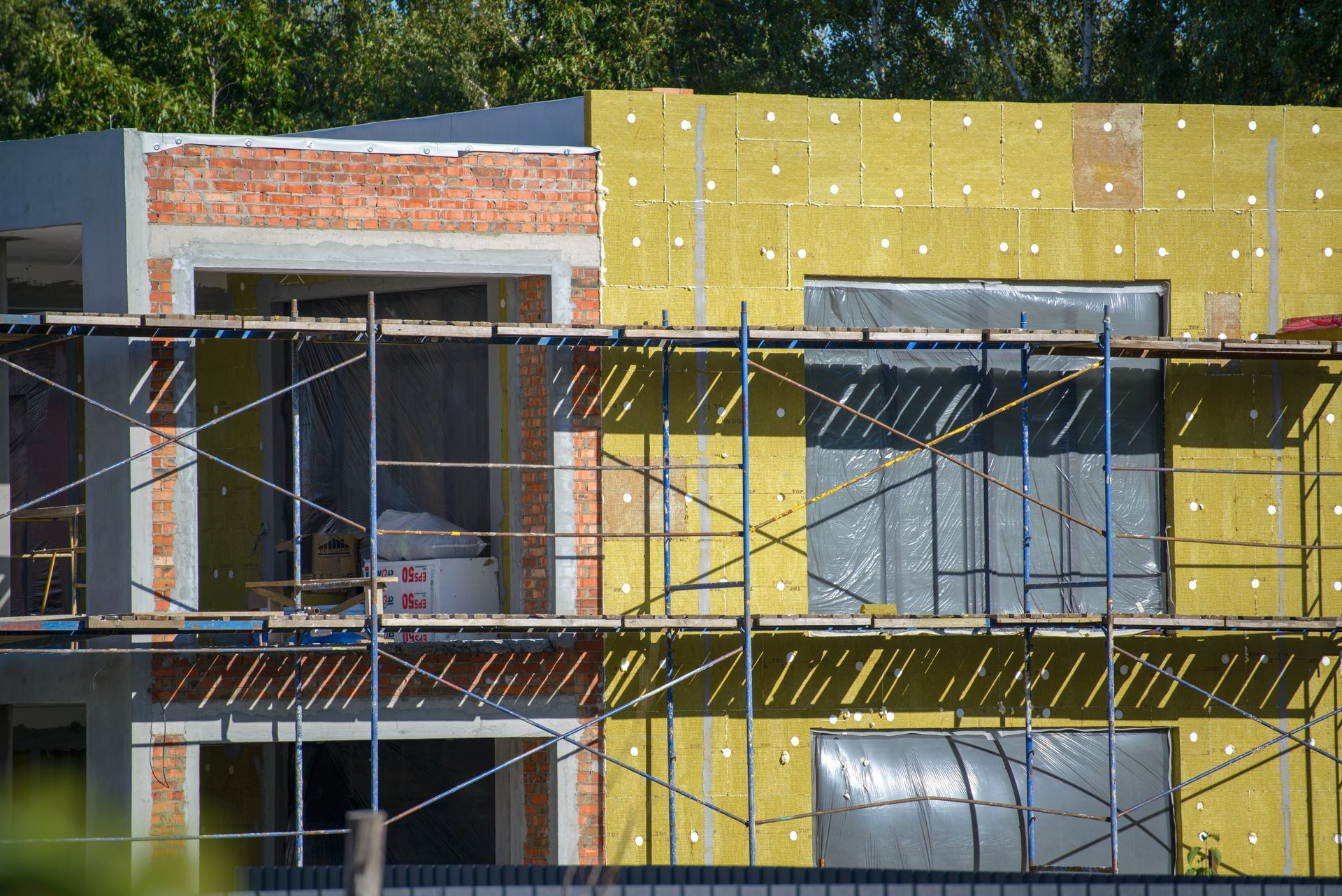 Façade d'un bâtiment partiellement construit, recouverte d'isolant jaune et d'échafaudages, avec de grandes fenêtres
