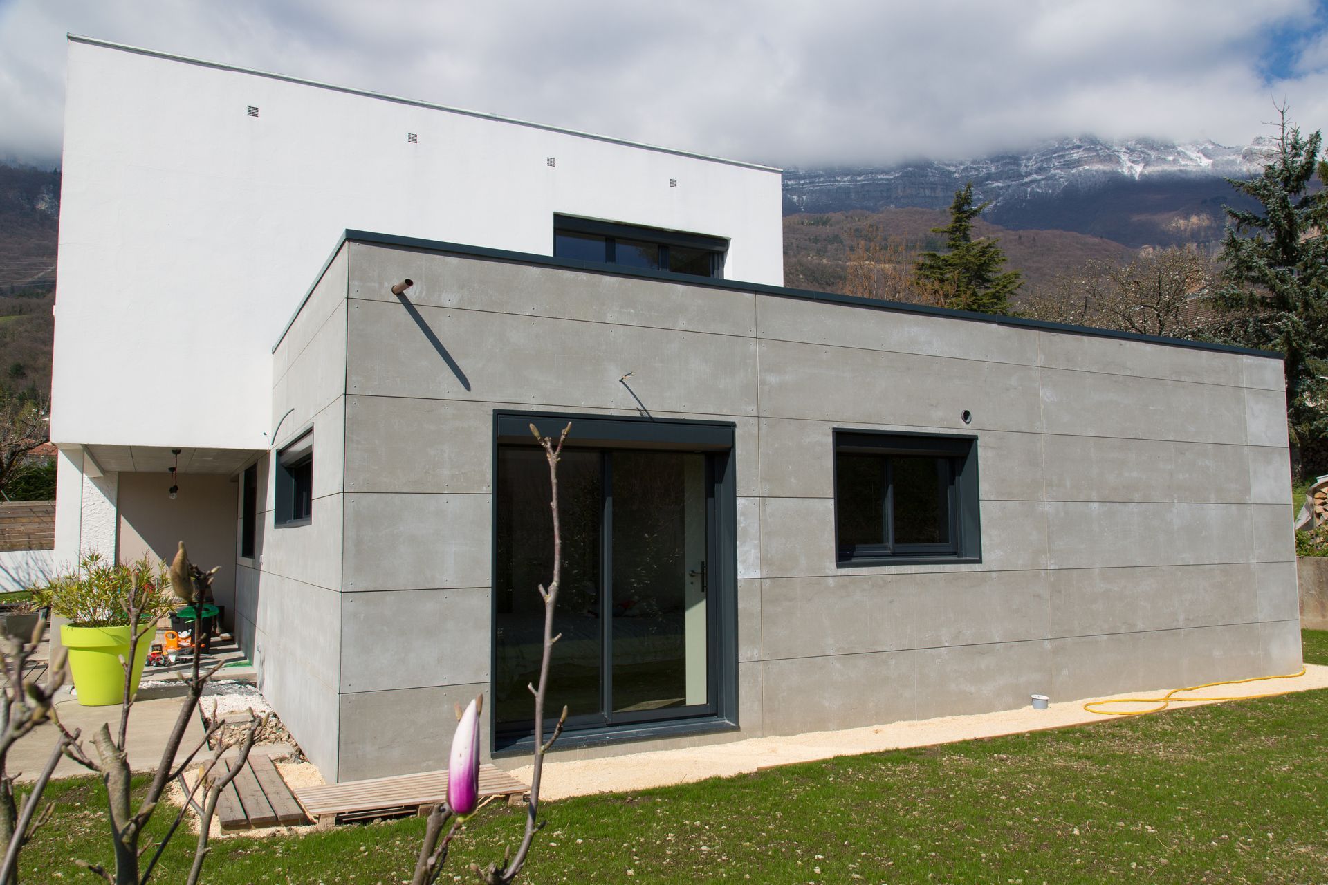 Extérieur d'une maison moderne grise et blanche avec une porte vitrée, une fenêtre et des montagnes en arrière-plan.