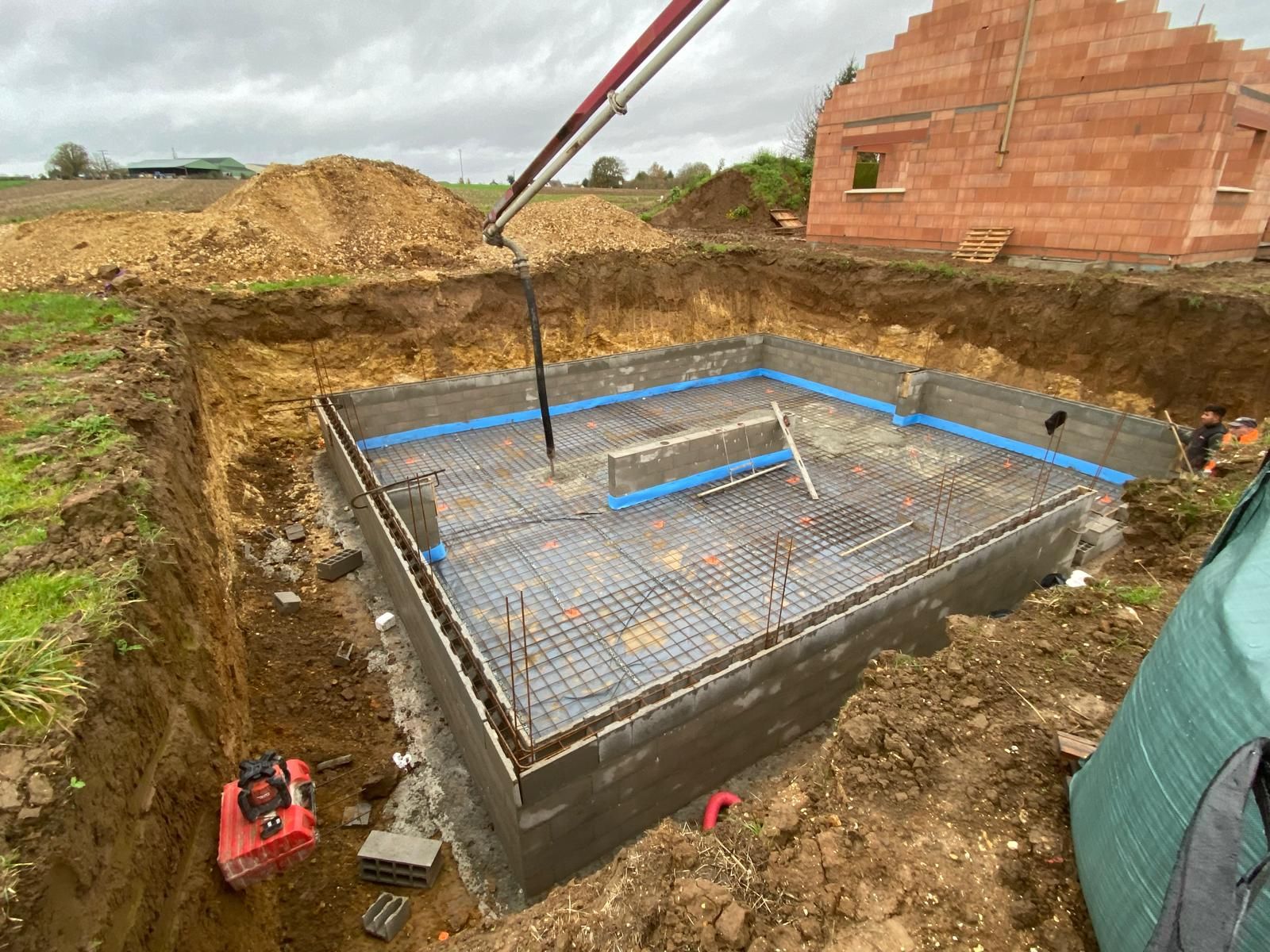 Fondation en béton en construction avec armatures, conduit bleu et tuyau de pompe dans une fosse d'excavation boueuse