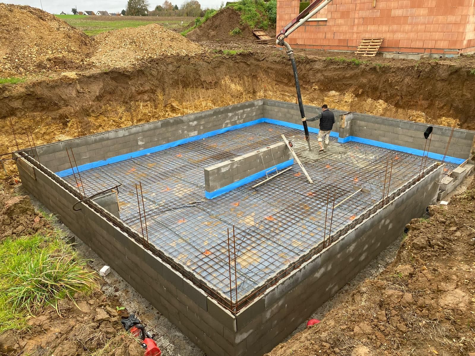 Semelle de fondation en armature métallique dans une excavation de terre, encadrée par des coffrages en béton et des repères de niveau bleus.
