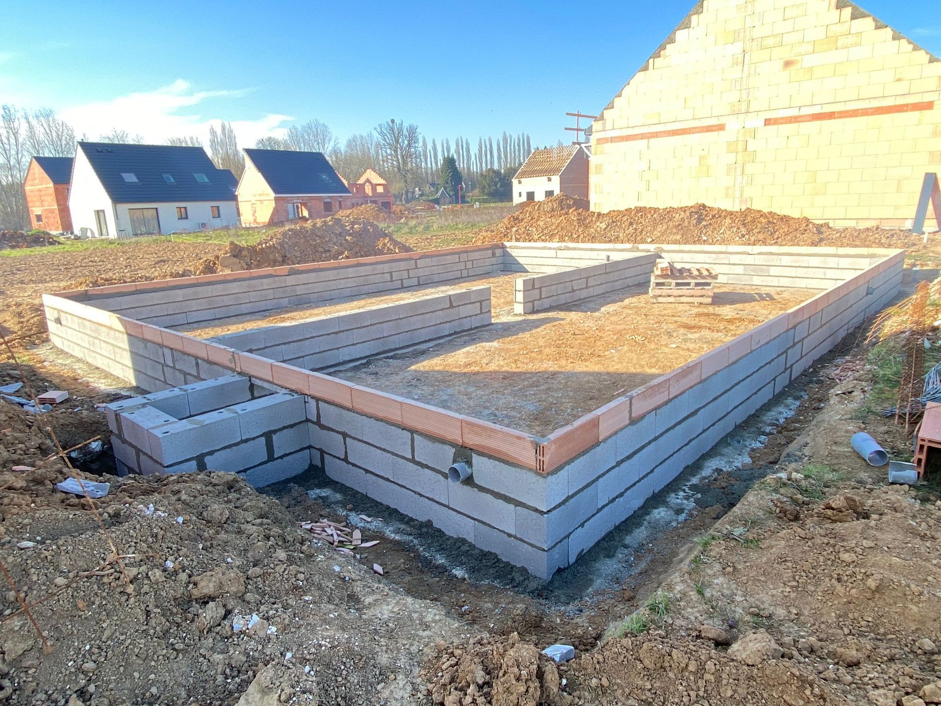 Murs de fondation d'une maison en construction sur un terrain vague, avec des maisons et un ciel bleu en arrière-plan.