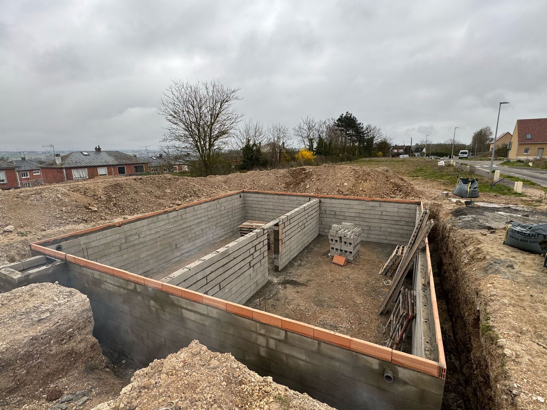 Fondations en béton partiellement construites avec des murs en parpaings sur un chantier boueux sous un ciel nuageux.