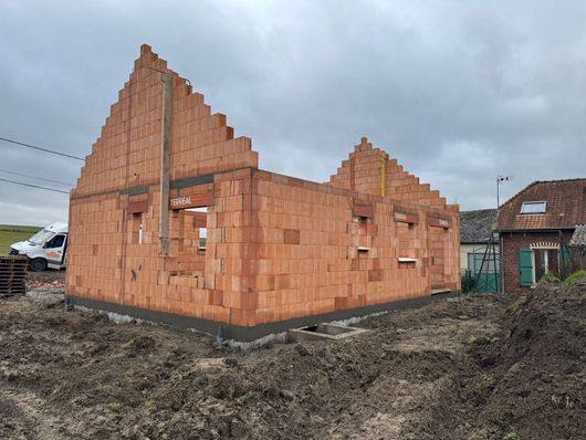 Maison en briques partiellement construite, avec un toit à pignons pentu, sur un chantier boueux sous un ciel nuageux.