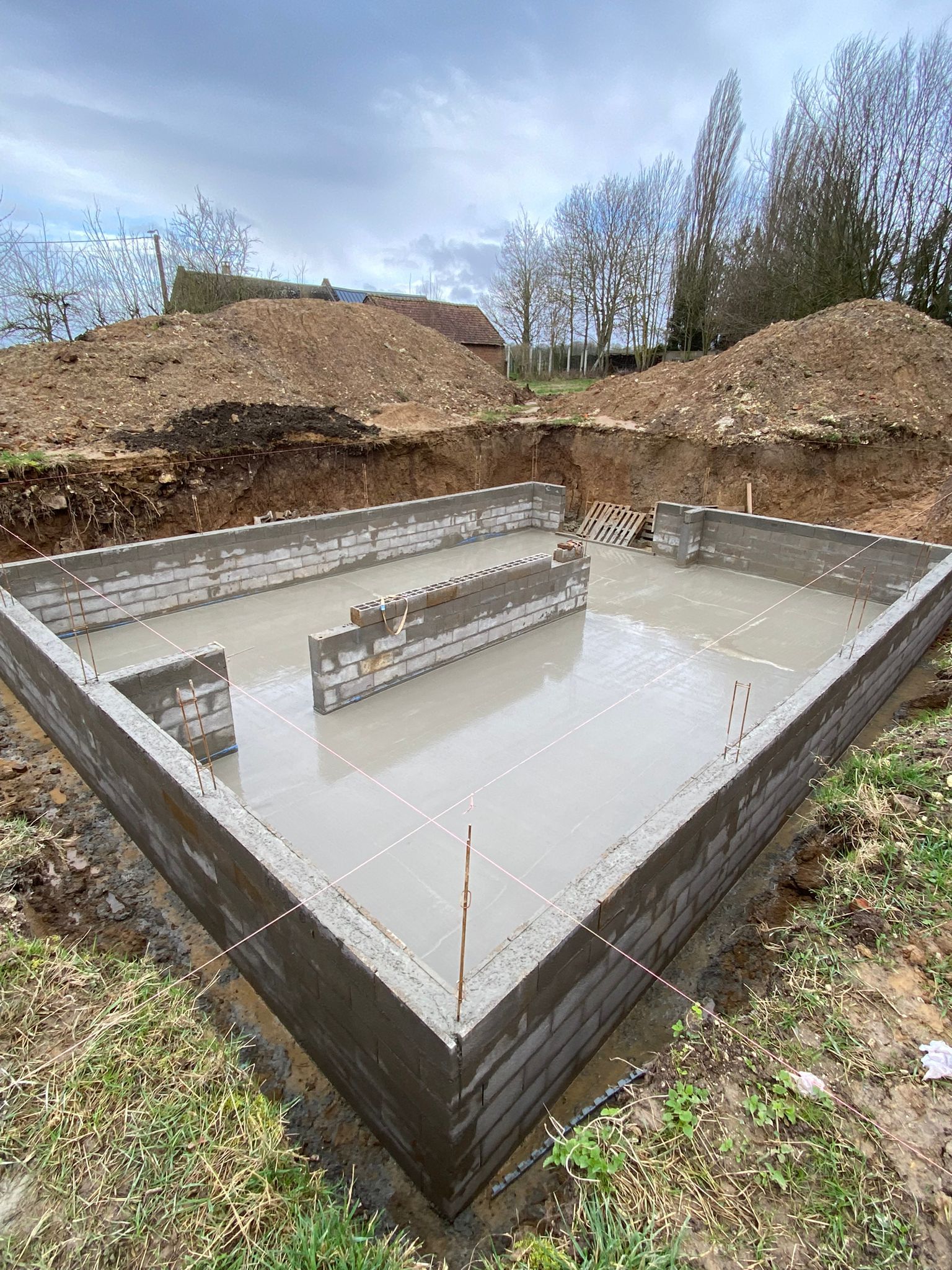 Dalle de fondation en béton avec murets dans un chantier de construction en excavation, entourée de tas de terre et d'arbres dénudés.