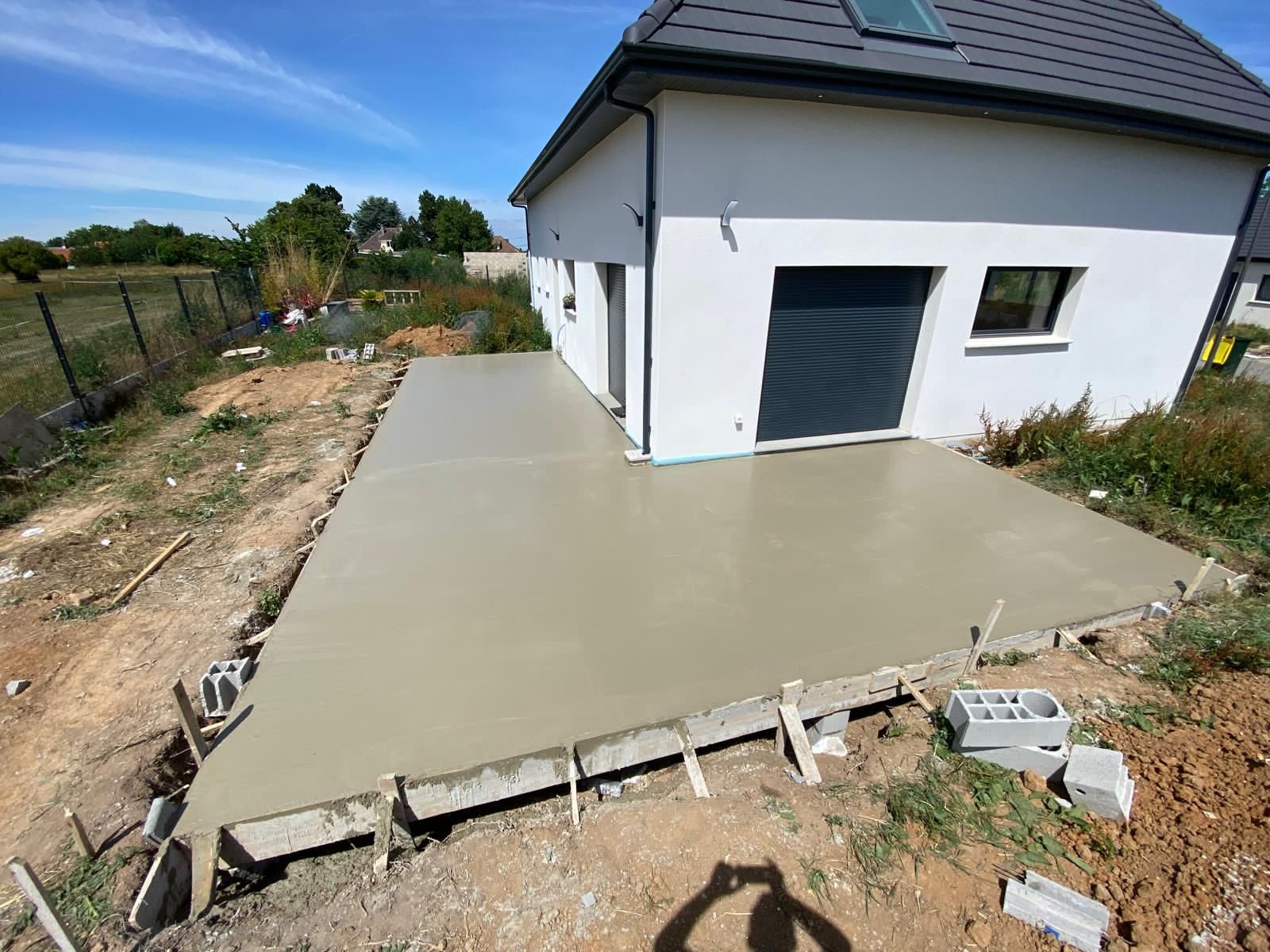 Dalle de fondation en béton en construction sur un terrain sablonneux avec des arbres et des maisons en arrière-plan.
