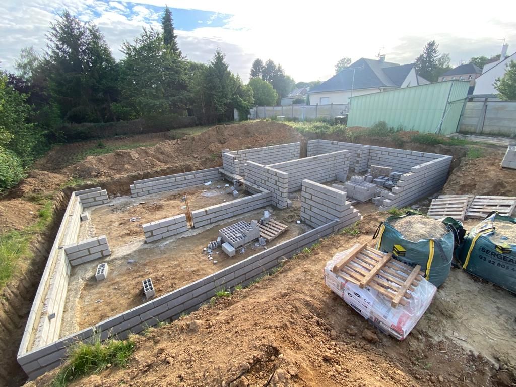 Chantier de construction avec murs de fondation et blocs de béton empilés sur un terrain en terre battue