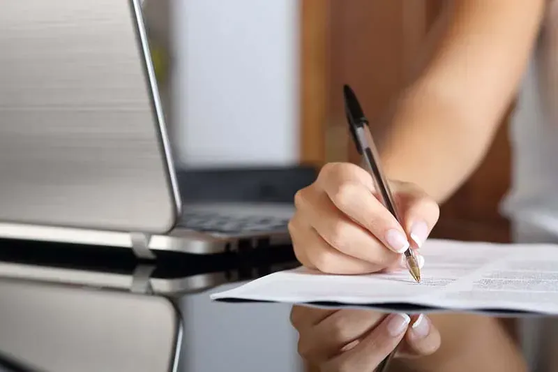 Una mujer está escribiendo en un trozo de papel frente a una computadora portátil.