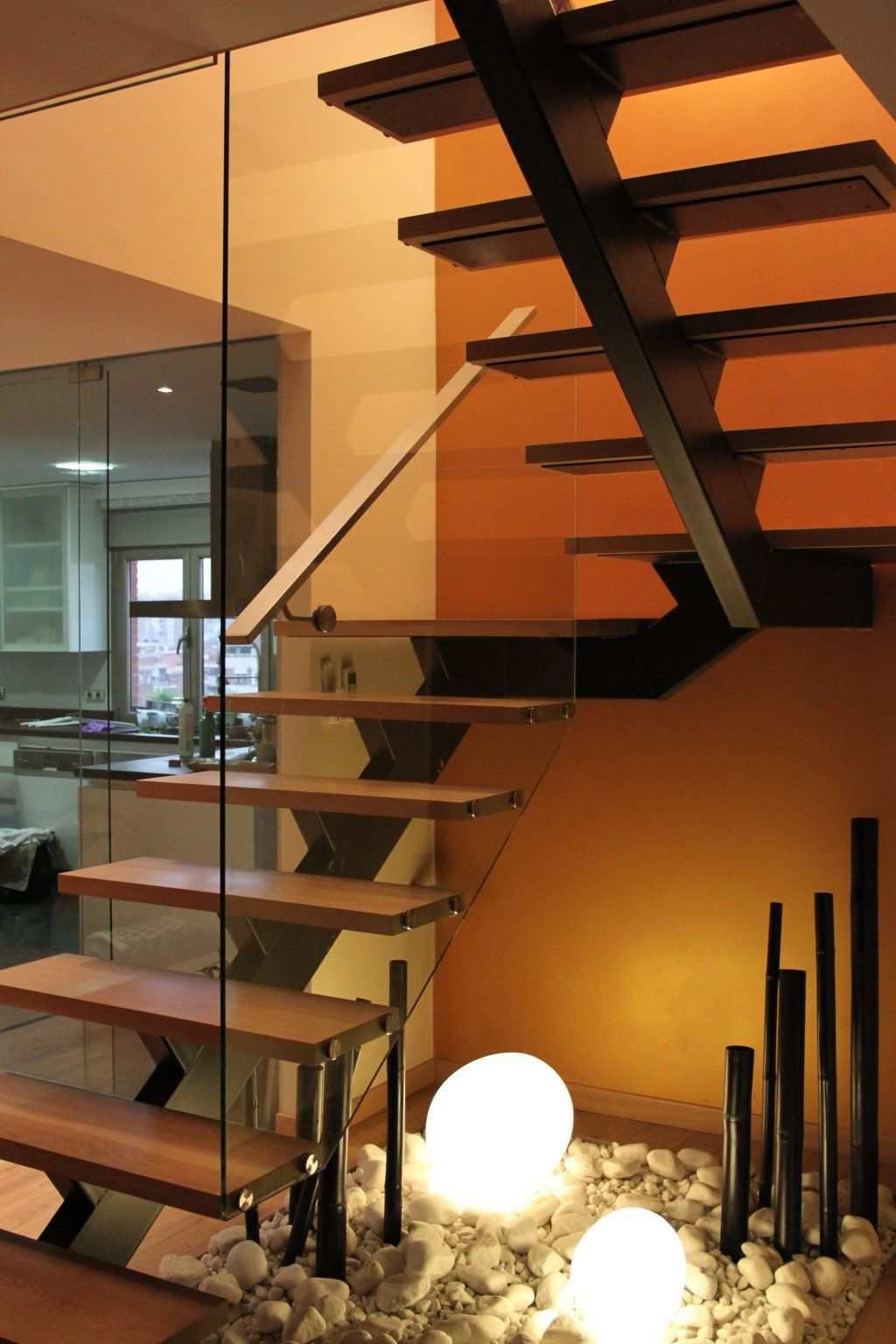 Escalera moderna con escalones de madera, barandilla de cristal y pared naranja con acento. Iluminada con luces decorativas.
