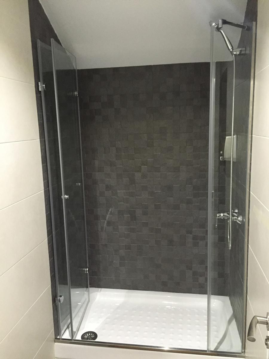 Mampara de ducha de cristal con pared de azulejos oscuros. Base de ducha blanca y paredes claras.