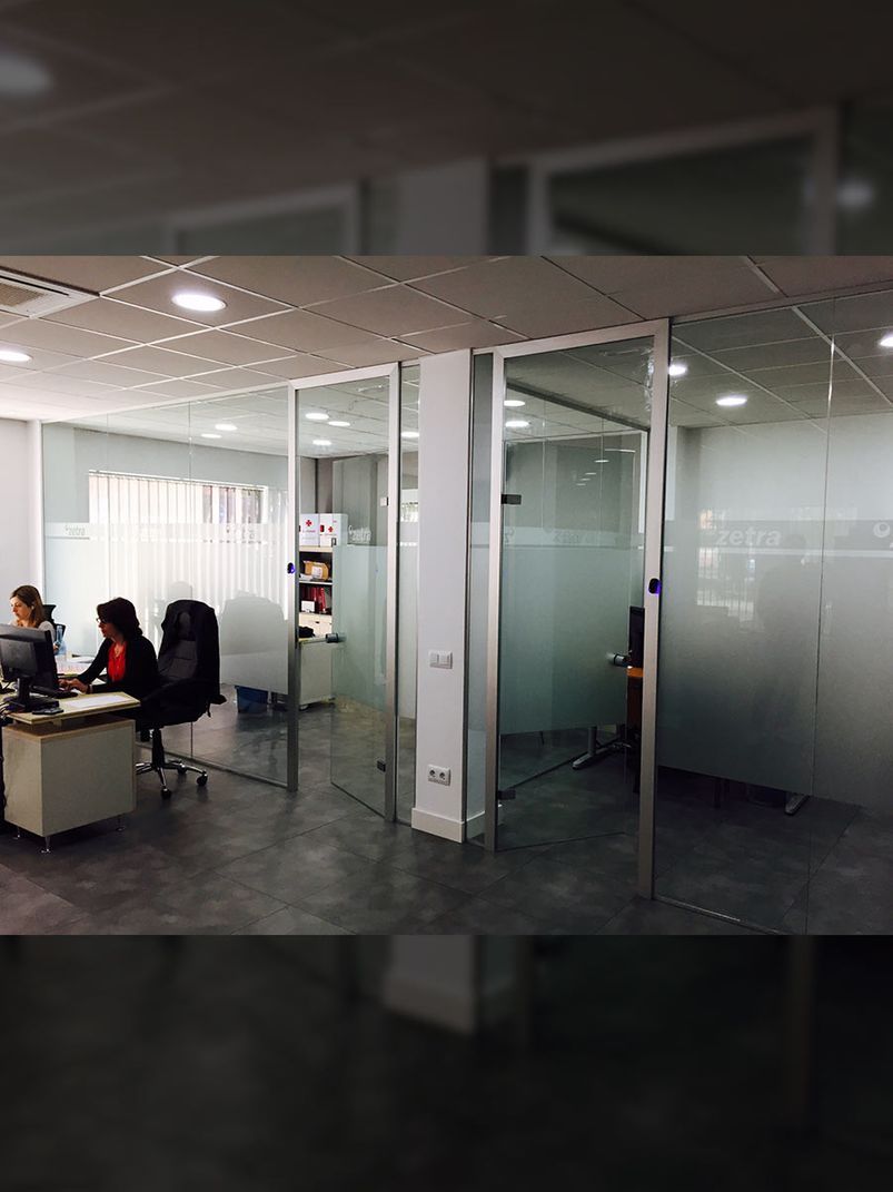Interior de oficina moderno con paredes de vidrio, personas trabajando en escritorios e iluminación empotrada.