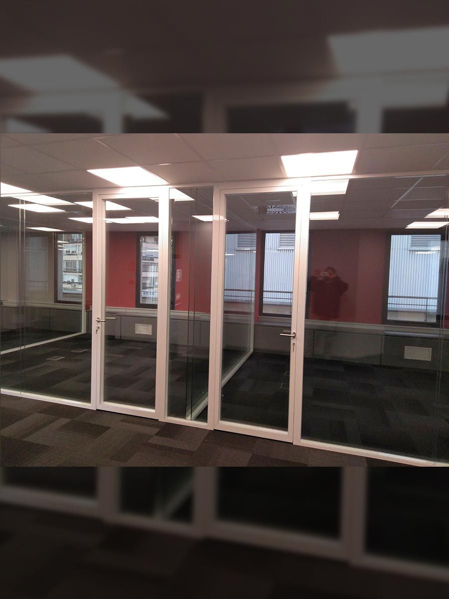 Pasillo de oficinas con habitaciones acristaladas. Ventanas con paneles rojos, alfombra gris y paredes blancas.