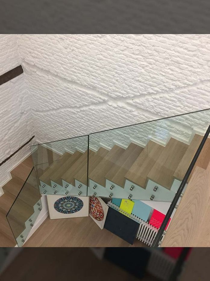 Escalera con peldaños de madera y barandilla de cristal, pared blanca texturizada. Cojines y muestras de color en la base.