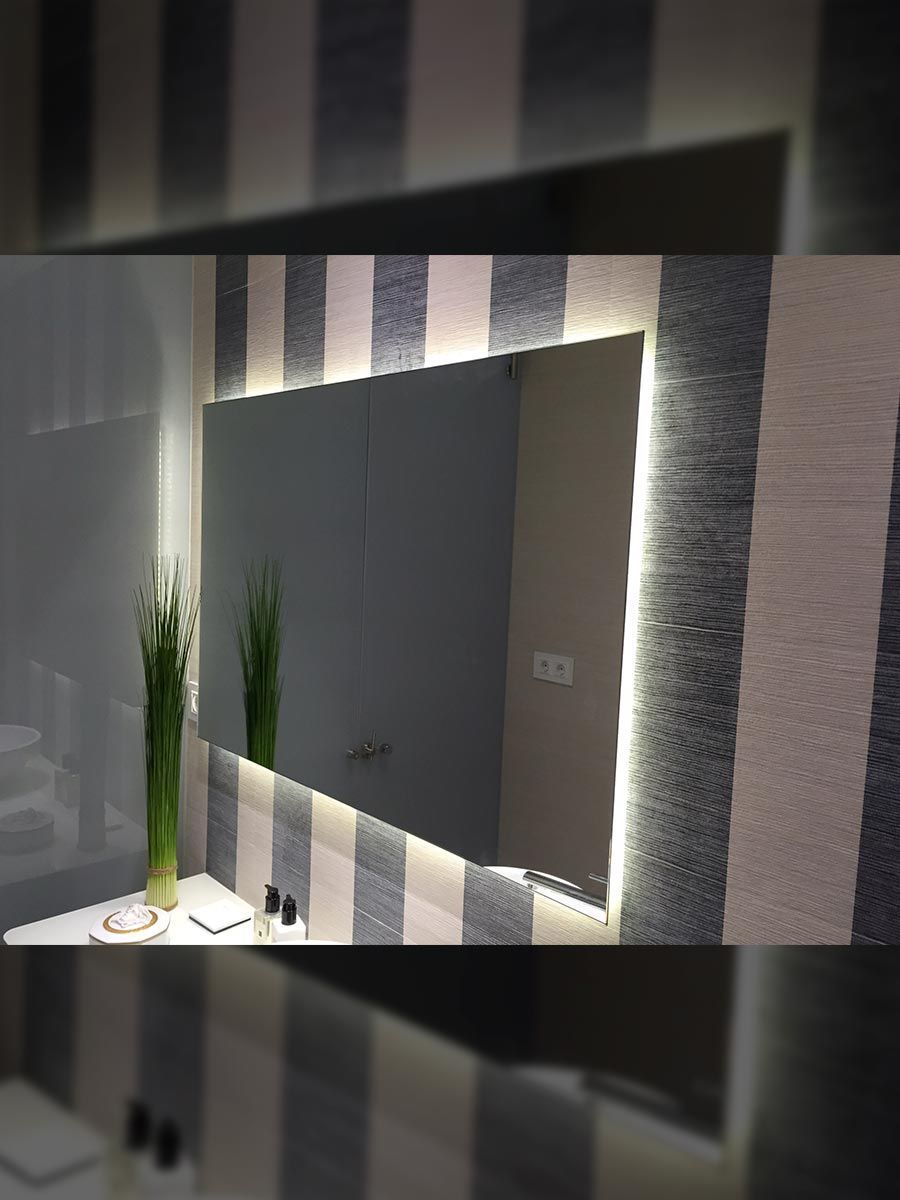 Un espejo grande y rectangular con iluminación LED, montado en una pared rayada sobre un mostrador blanco con decoración.