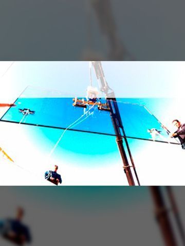 Un gran panel de vidrio siendo levantado por una grúa, con trabajadores cerca bajo un cielo azul brillante.