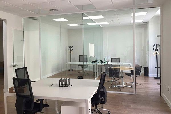 Oficina moderna con paredes de vidrio, escritorios blancos, sillas y pisos de madera.