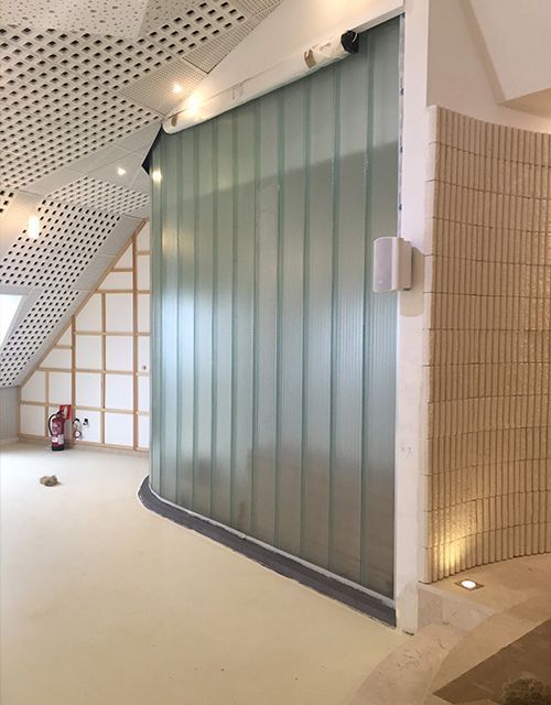 La pared de ladrillos de vidrio se curva entre paredes beige, un piso claro y un techo blanco perforado.