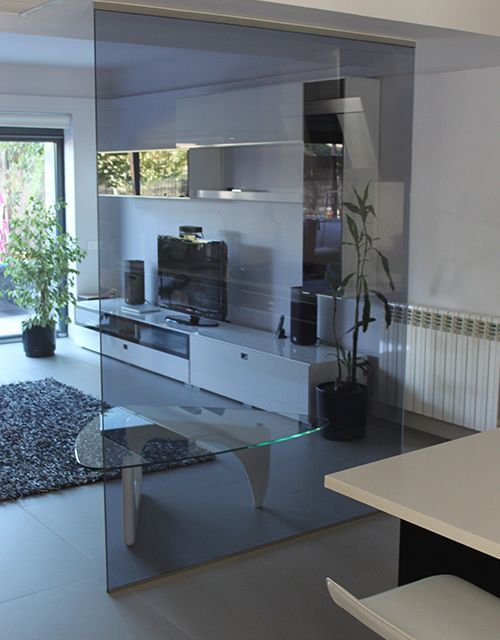 Biombo de vidrio que oculta parcialmente un espacio de estar moderno con muebles blancos, TV y plantas.