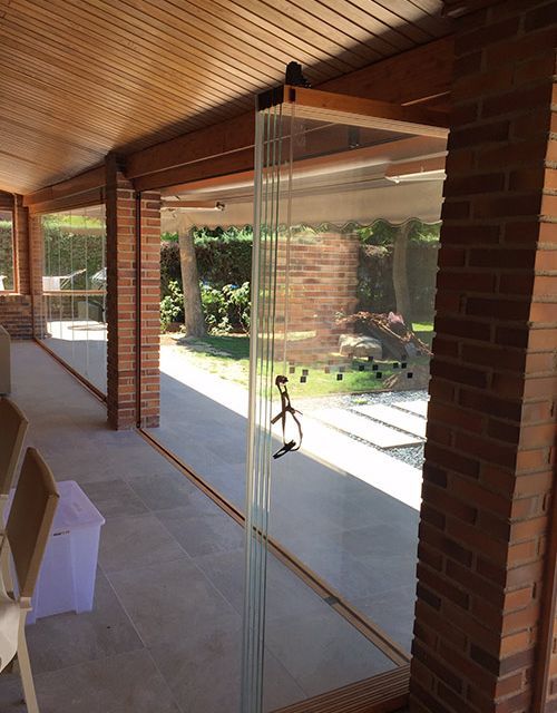 Grandes paneles de vidrio forman una pared en un patio con columnas de ladrillo y techo de madera.