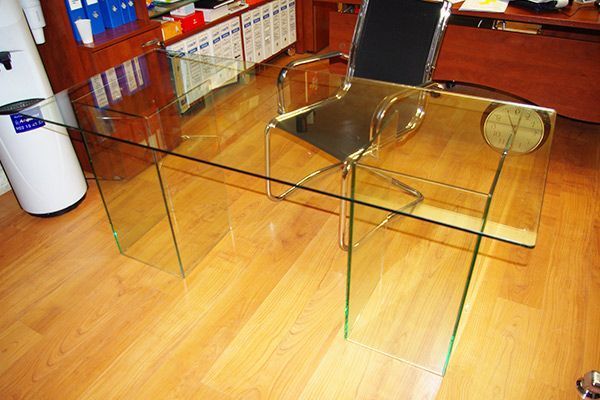 Escritorio de vidrio en oficina con silla transparente, piso de madera y dispensador de agua.