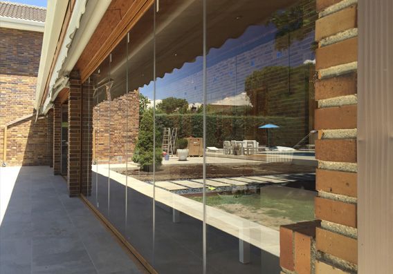 Patio con paneles de vidrio transparente que reflejan el patio trasero, junto a una pared de ladrillo y un toldo de madera.