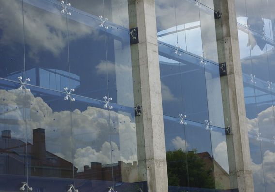 Fachada de vidrio que refleja el cielo azul con nubes, fijada a columnas de hormigón mediante herrajes metálicos de araña.