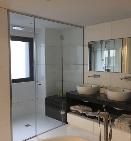 Baño blanco moderno con ducha de cristal, lavabos dobles sobre tocador oscuro y una ventana.