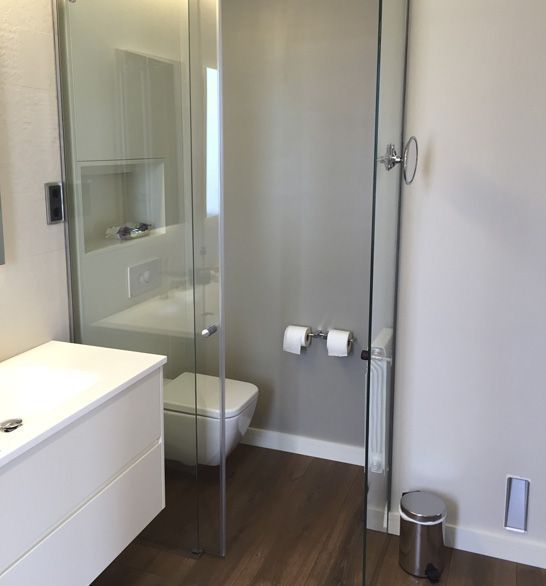 Baño moderno con grifería blanca, ducha de cristal y suelo imitación madera. Paredes grises, inodoro y un pequeño cubo de basura.