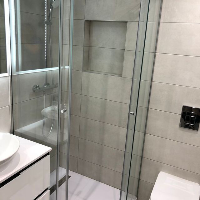 Cabina de ducha con puertas de cristal, azulejos blancos y estante empotrado.