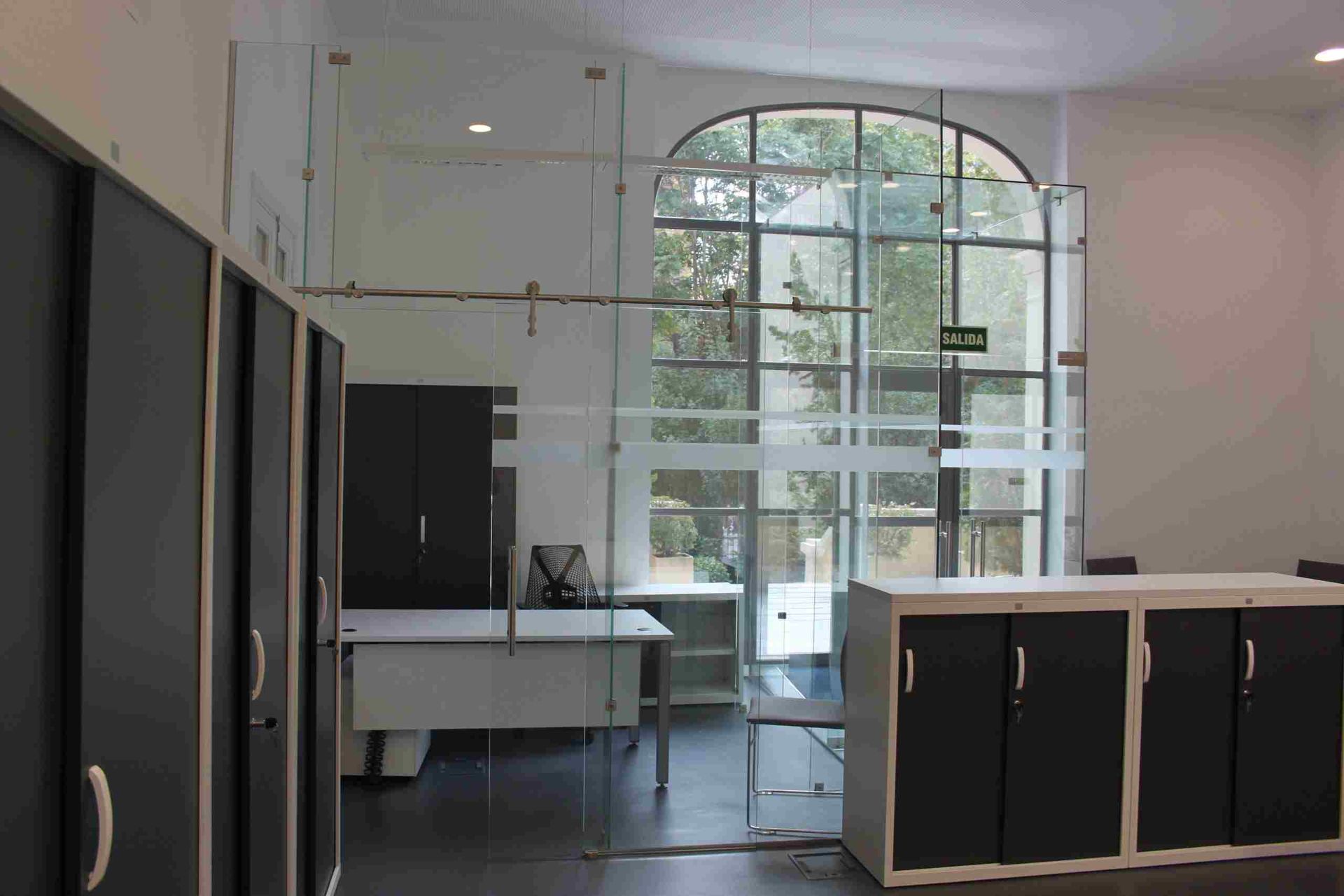 Interior de oficina con paredes de vidrio, gabinetes y escritorio cerca de una ventana arqueada con vista a la vegetación.