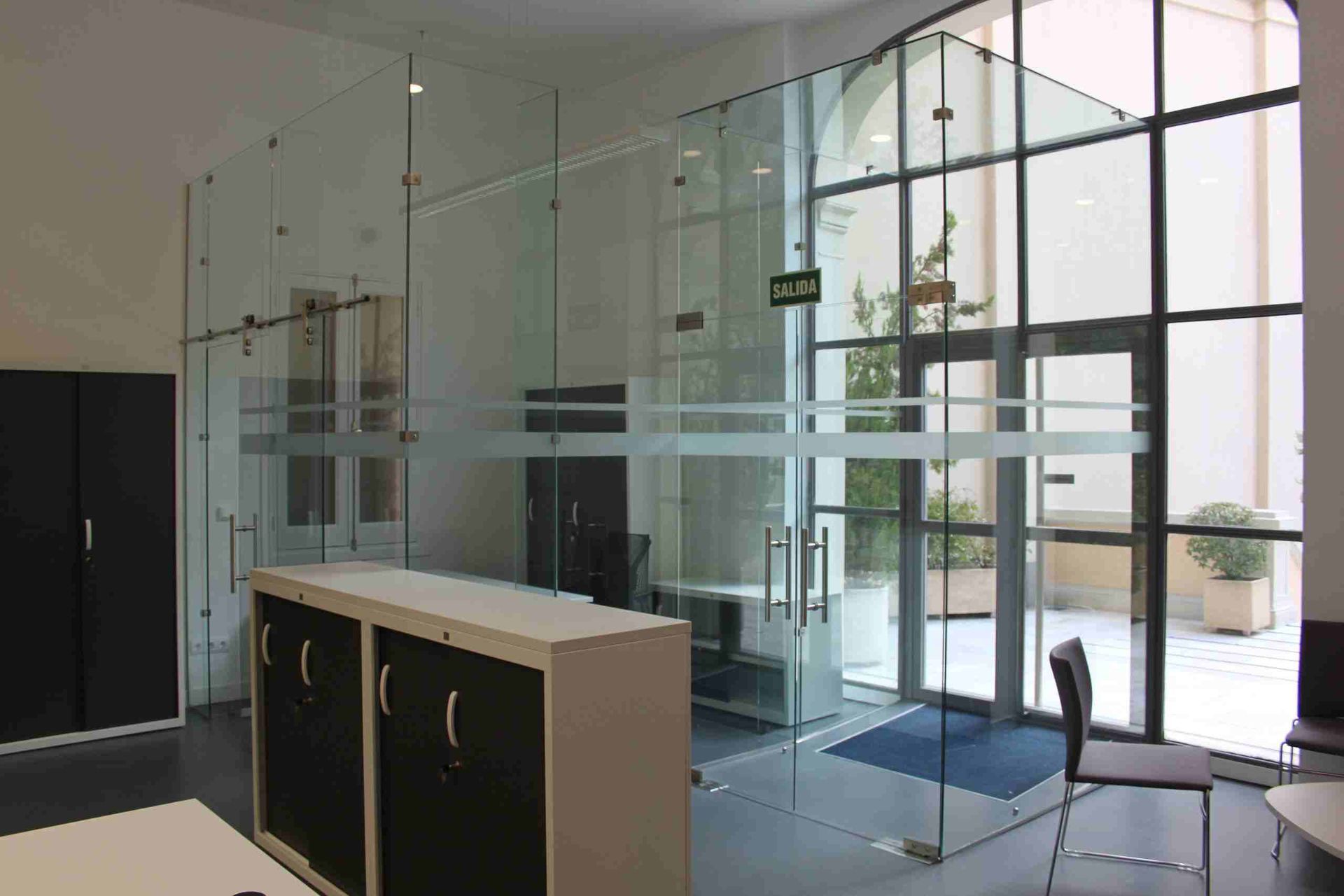 Entrada de oficina con paredes de cristal, armario y señal de salida. La puerta da al exterior.