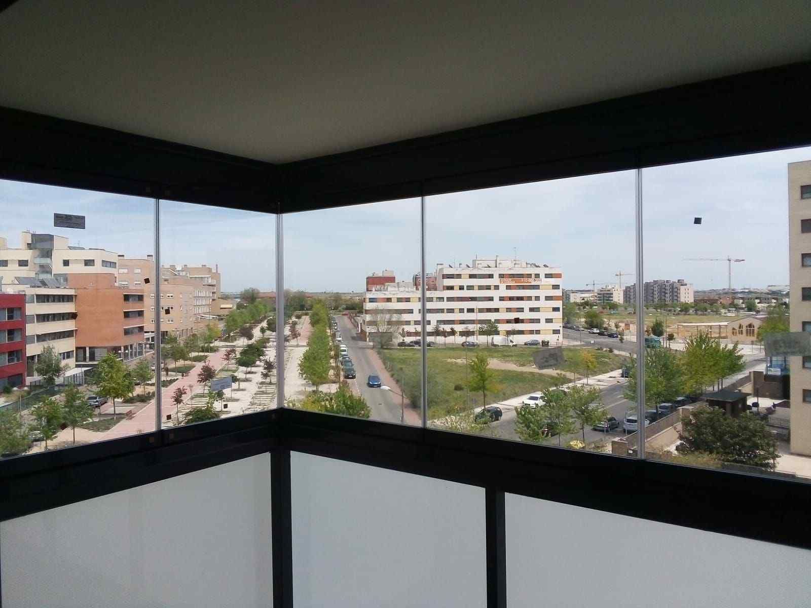 Vista desde un balcón con paredes de vidrio con vista a una calle de la ciudad bordeada de árboles y edificios bajo un cielo brillante.