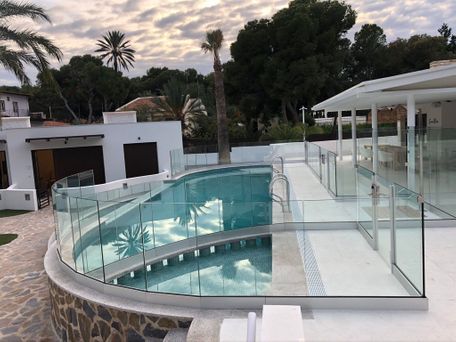 Piscina rodeada de barandilla de cristal en un patio blanco. Palmeras al fondo.