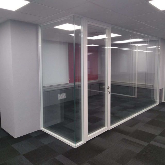 Oficina con paredes de cristal, marcos blancos, escritorios visibles y alfombra oscura.