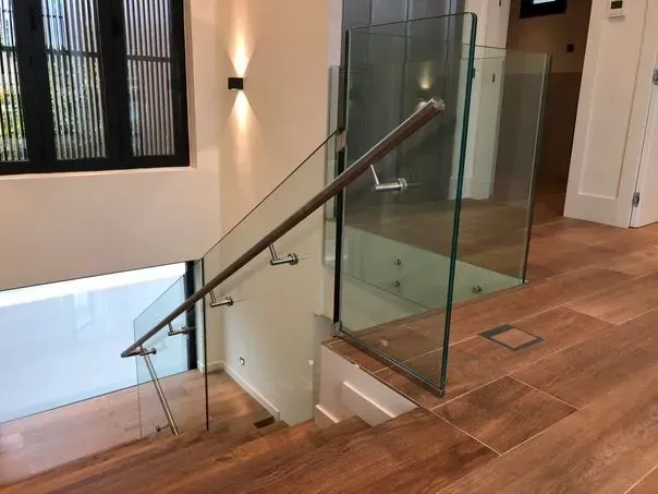 Escalera con barandilla de cristal y peldaños de madera, con suelo marrón y ventana.