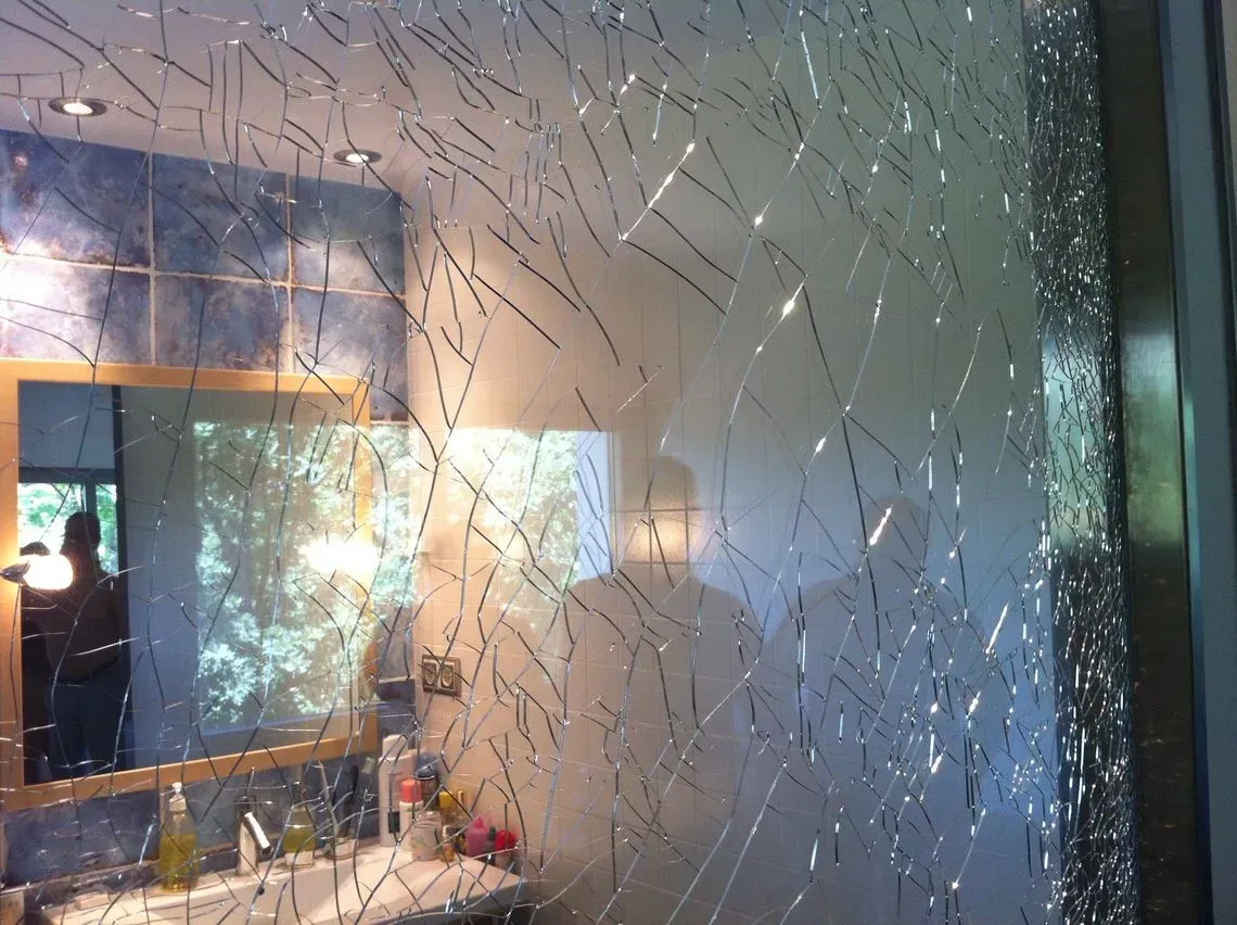 La pared de vidrio rota revela el interior de un baño con espejo y lavabo.
