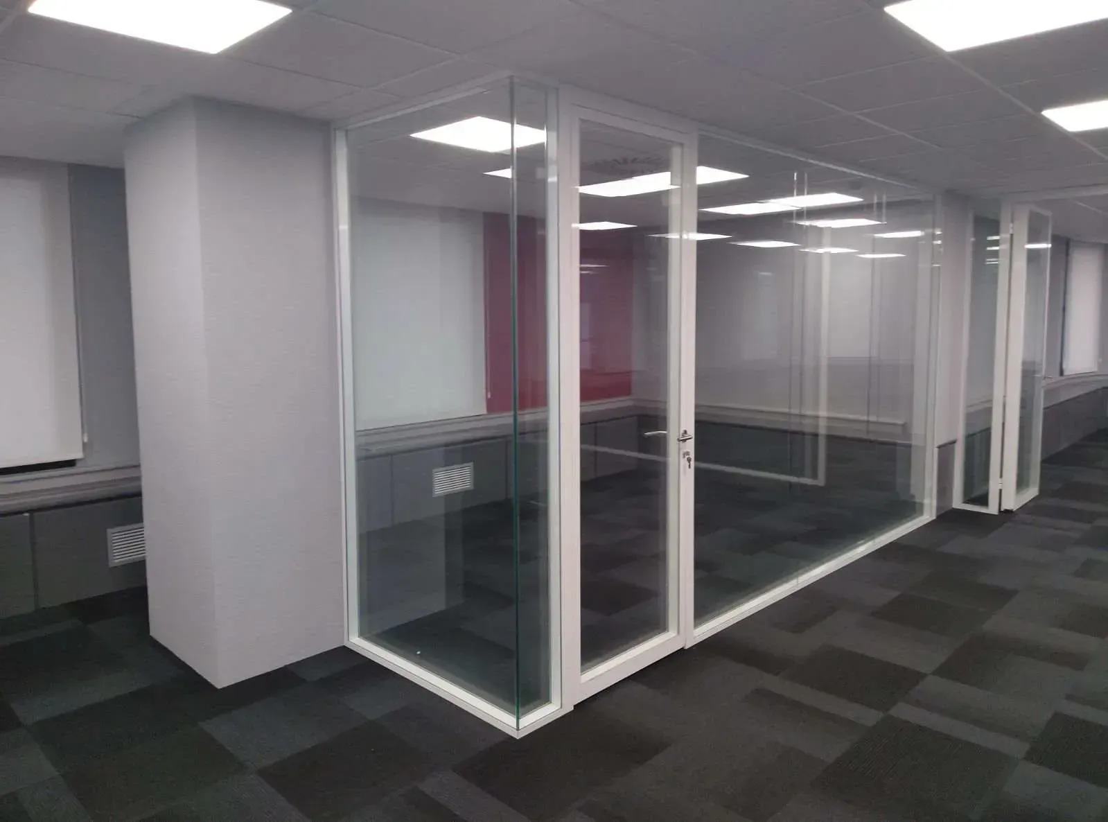 Oficina con cubículos con paredes de vidrio, marcos blancos y alfombra oscura; algunas estaciones de trabajo visibles.