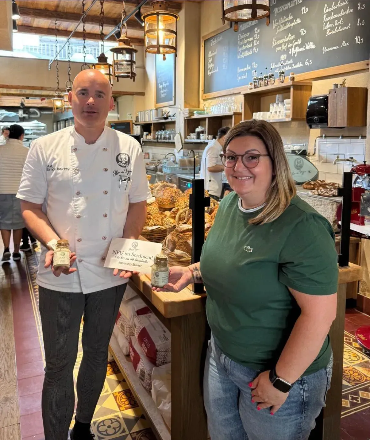 Mitarbeiter in der Bäckerei, die Einmachgläser halten und lächeln. Im Hintergrund hölzerne Theke und Regale.