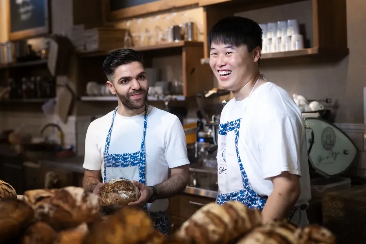 Zwei Bäcker in Schürzen lächeln in einer Bäckerei und halten Brot in den Händen. 