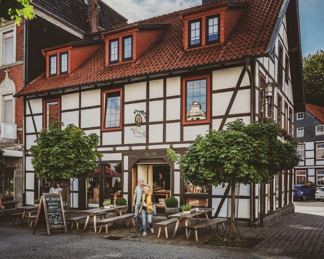 Fachwerkhaus mit rotem Dach; Sitzgelegenheiten im Freien. Passanten in der Nähe des Eingangs.