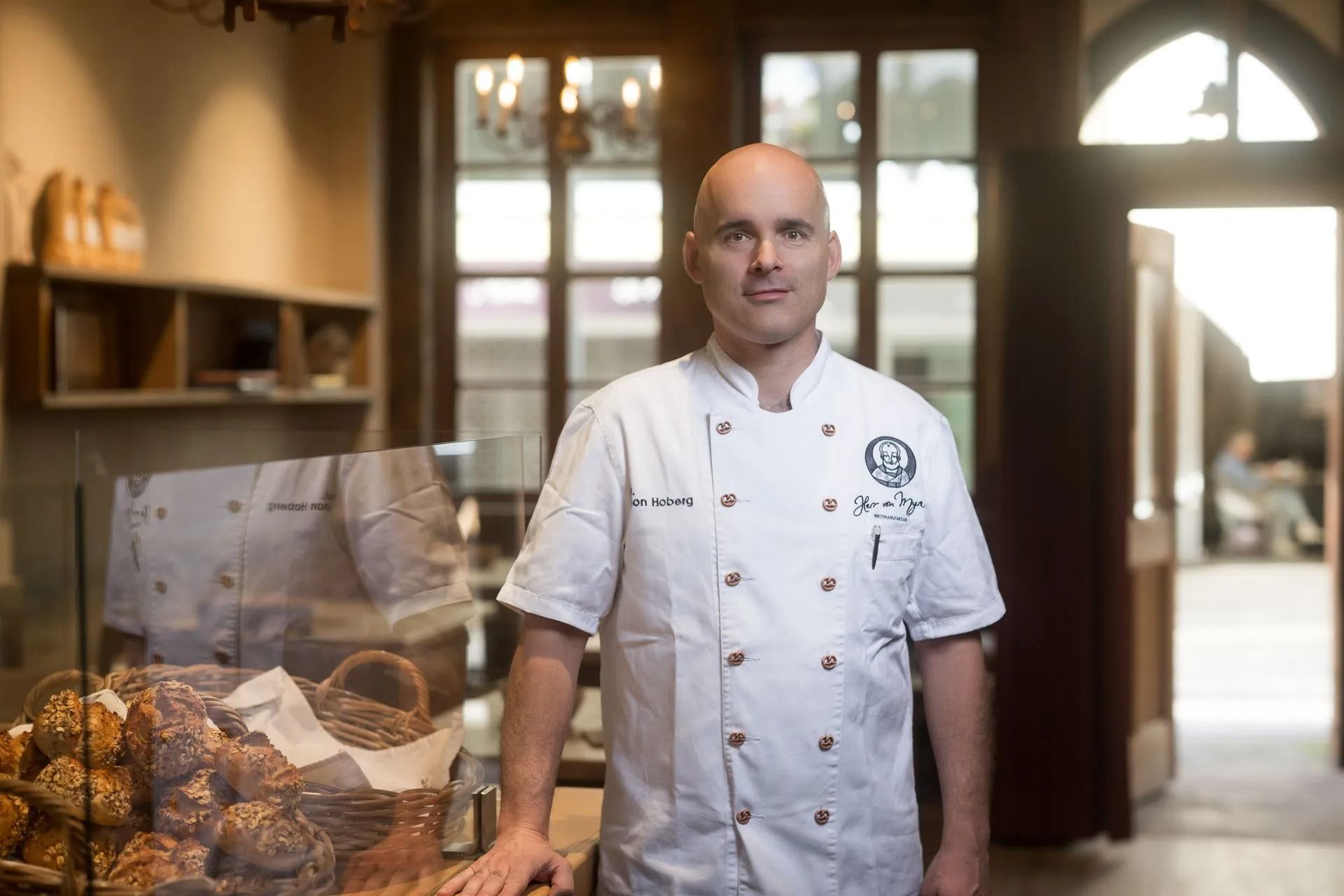 Ein Bäcker in weißer Uniform steht an der Theke einer Bäckerei und blickt in die Kamera.