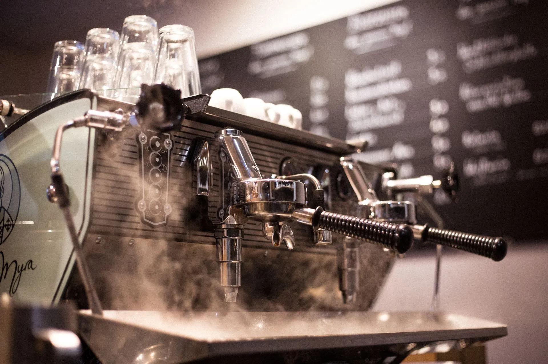 Espressomaschine in einem Café, dampfend. Chrom und Mintgrün, darüber gestapelte Gläser.