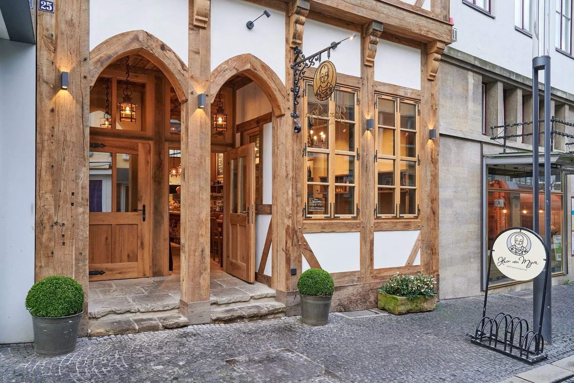 Außenansicht der Bäckerei mit Bogentüren und großen Fenstern. Steinpflaster, Pflanzen in Töpfen.