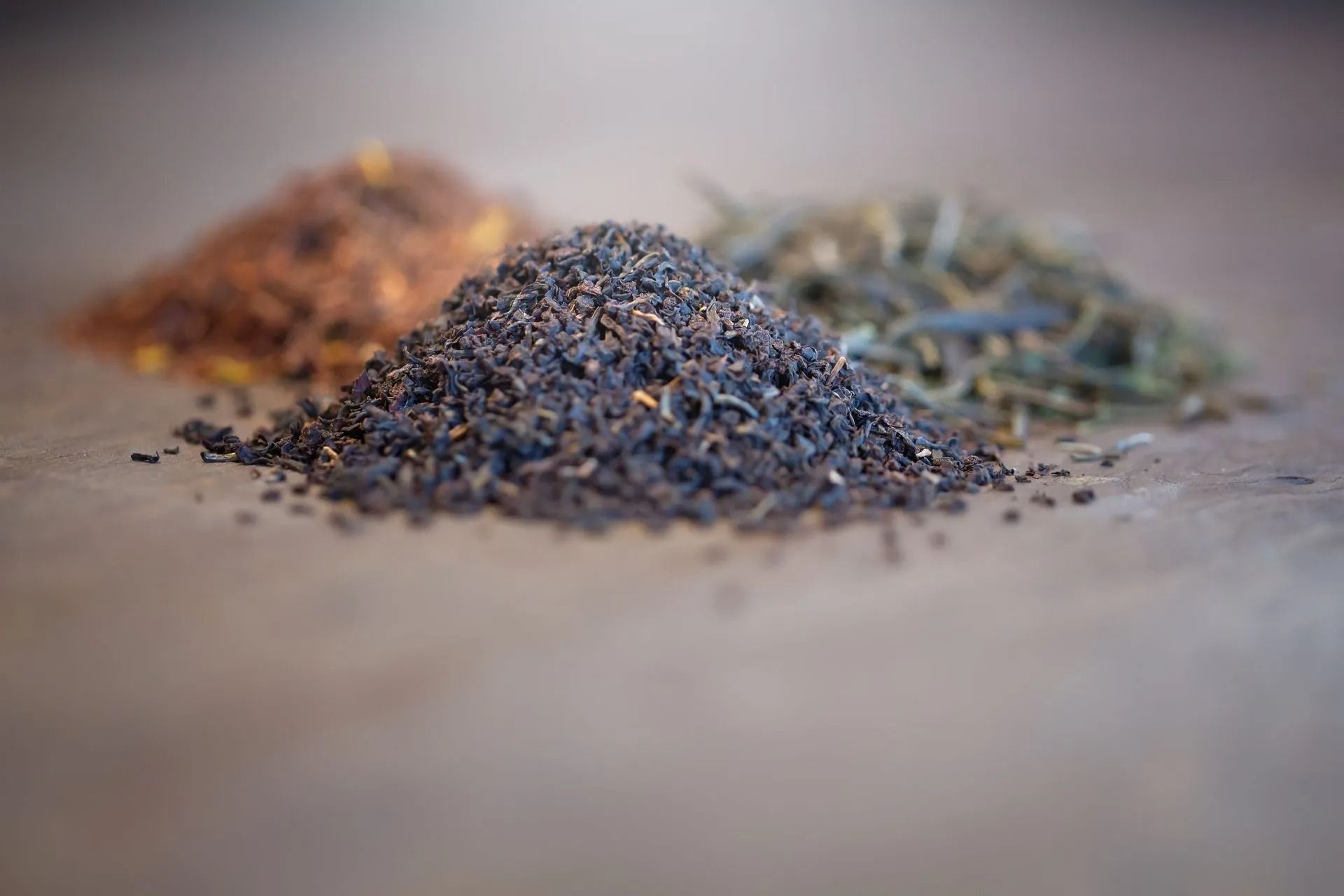 Drei Haufen Teeblätter: schwarze, rotbraune und grüne, angeordnet auf einer strukturierten Oberfläche.