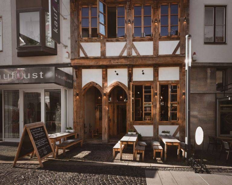 Fachwerkgebäude mit gewölbtem Eingang, Sitzgelegenheiten im Freien und einer handgeschriebenen Speisekarte.