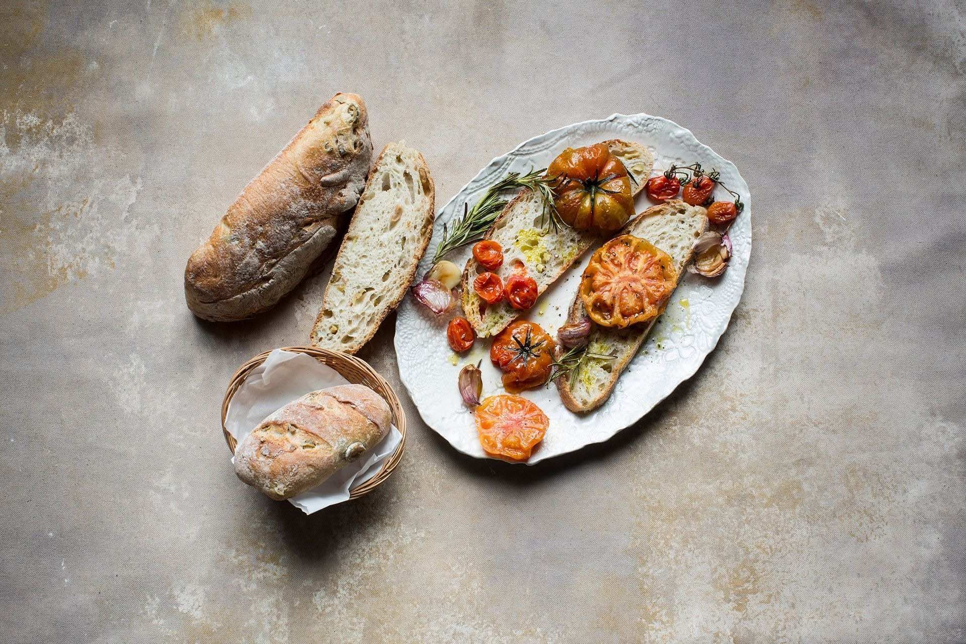 Brot und geröstete Tomaten auf einem Teller, daneben geschnittenes Brot und ein Laib in einem Korb.