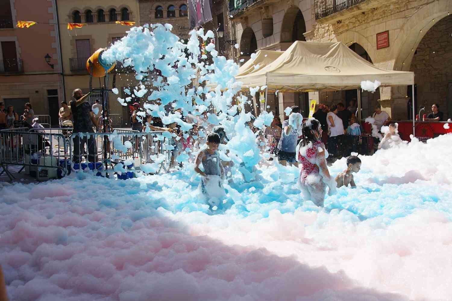 Gente jugando en una plaza llena de espuma rosa y azul, con edificios y tiendas de campaña al fondo.
