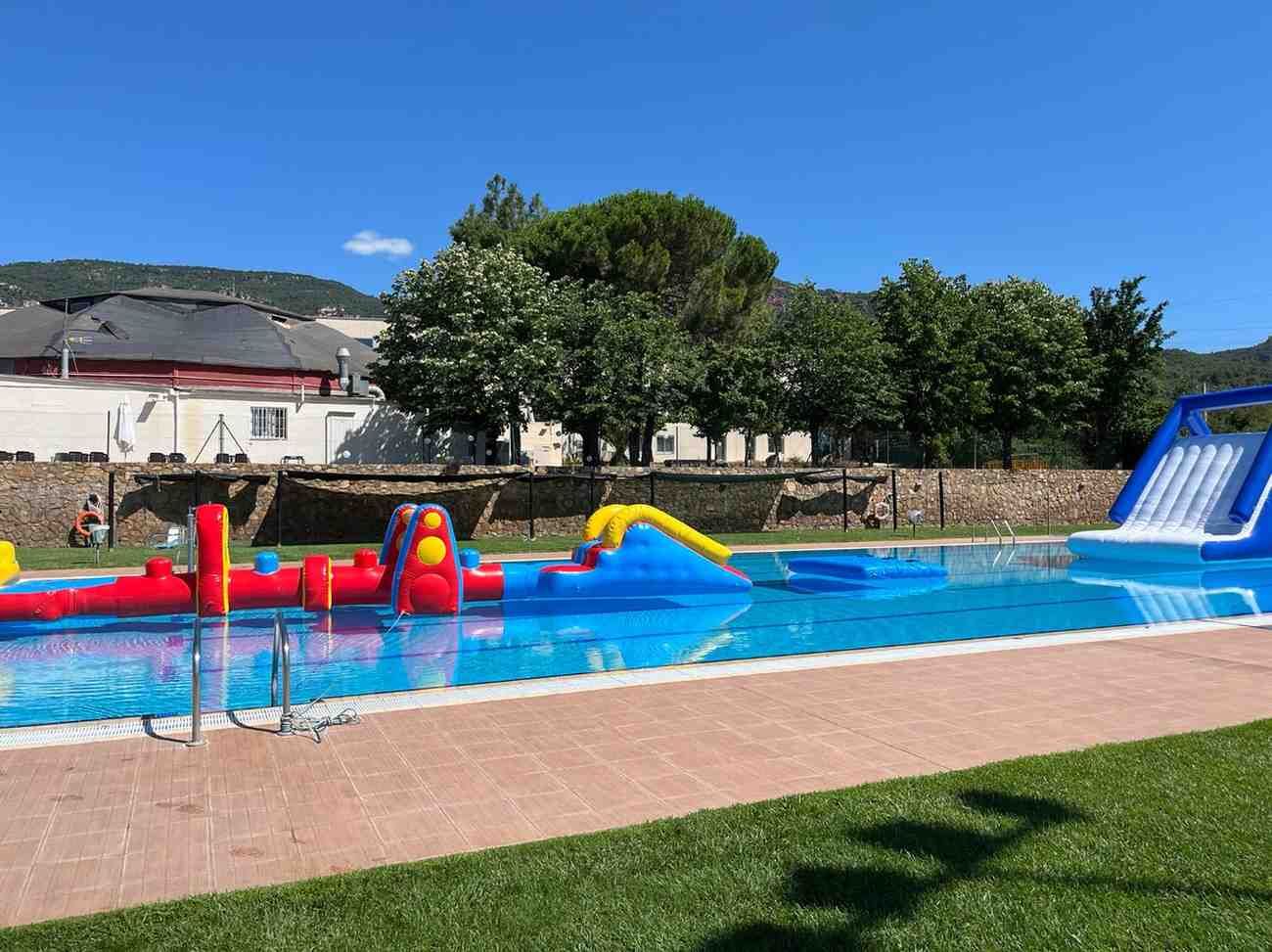 Piscina al aire libre con coloridos obstáculos inflables de agua y un tobogán azul en un día soleado.