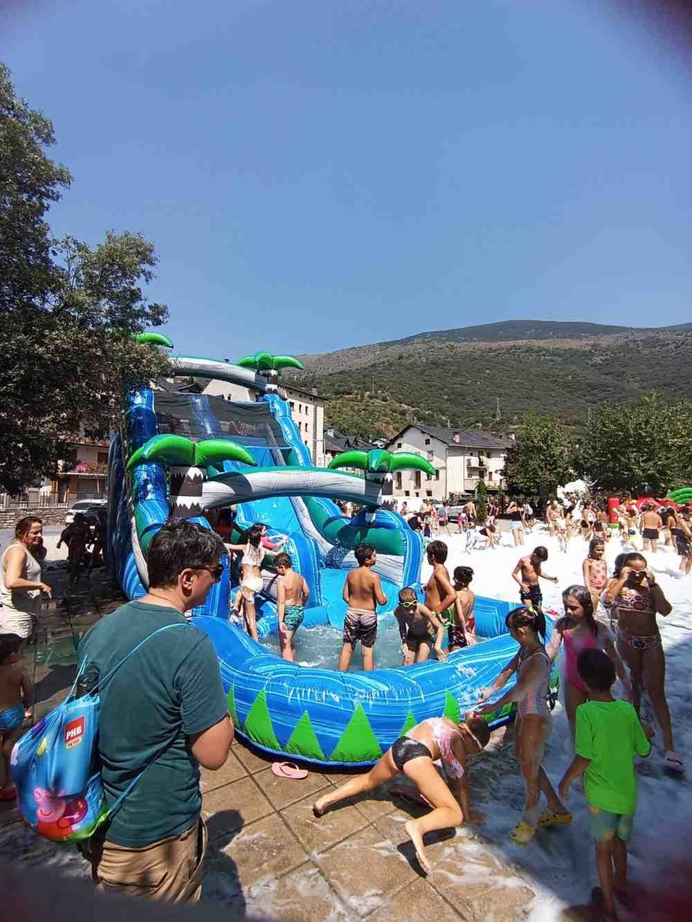 Niños jugando en una piscina inflable azul verdosa con tobogán en un parque acuático al aire libre.