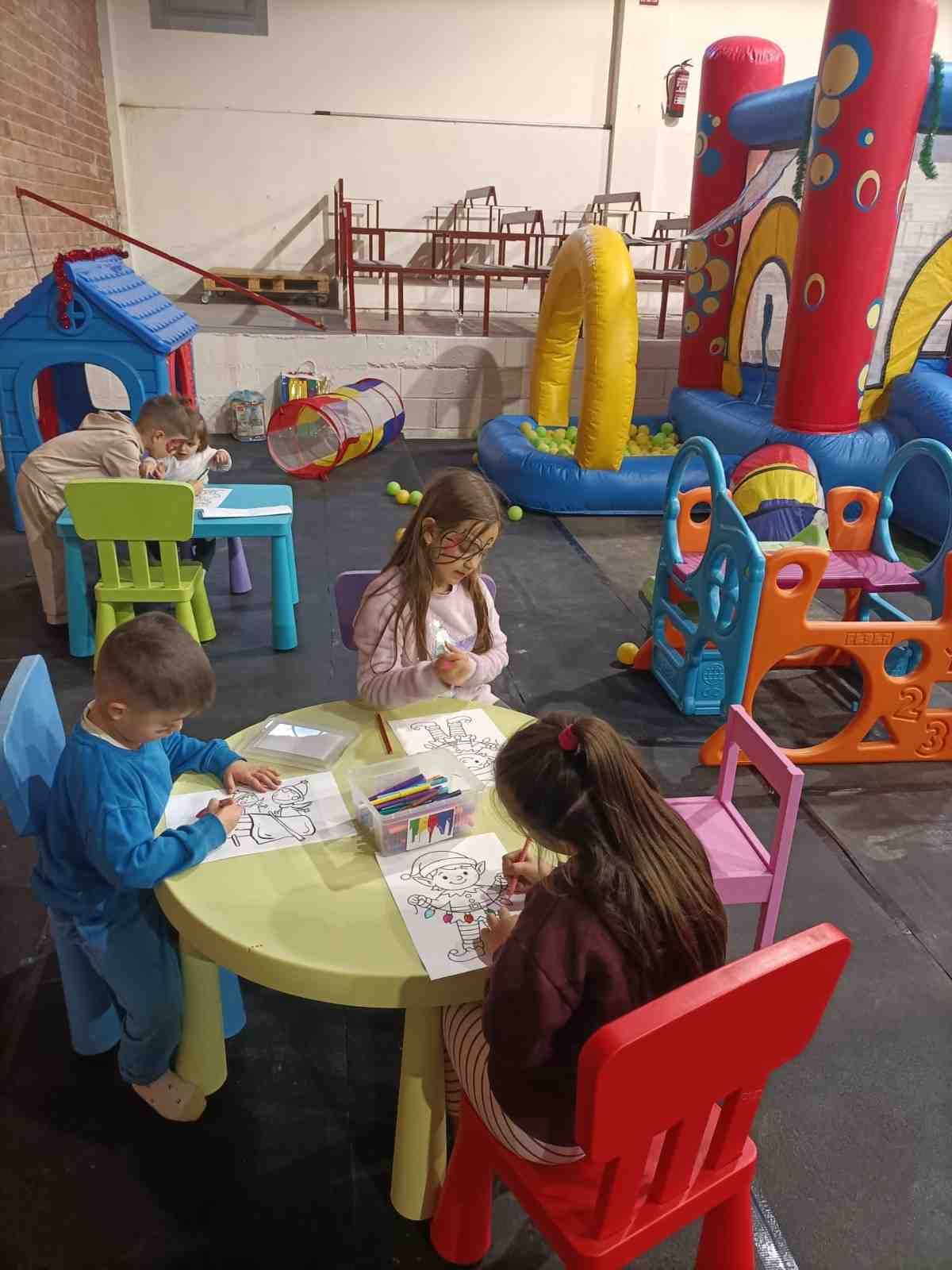 Niños coloreando en una mesa redonda en una sala de juegos con juguetes coloridos y un tobogán.